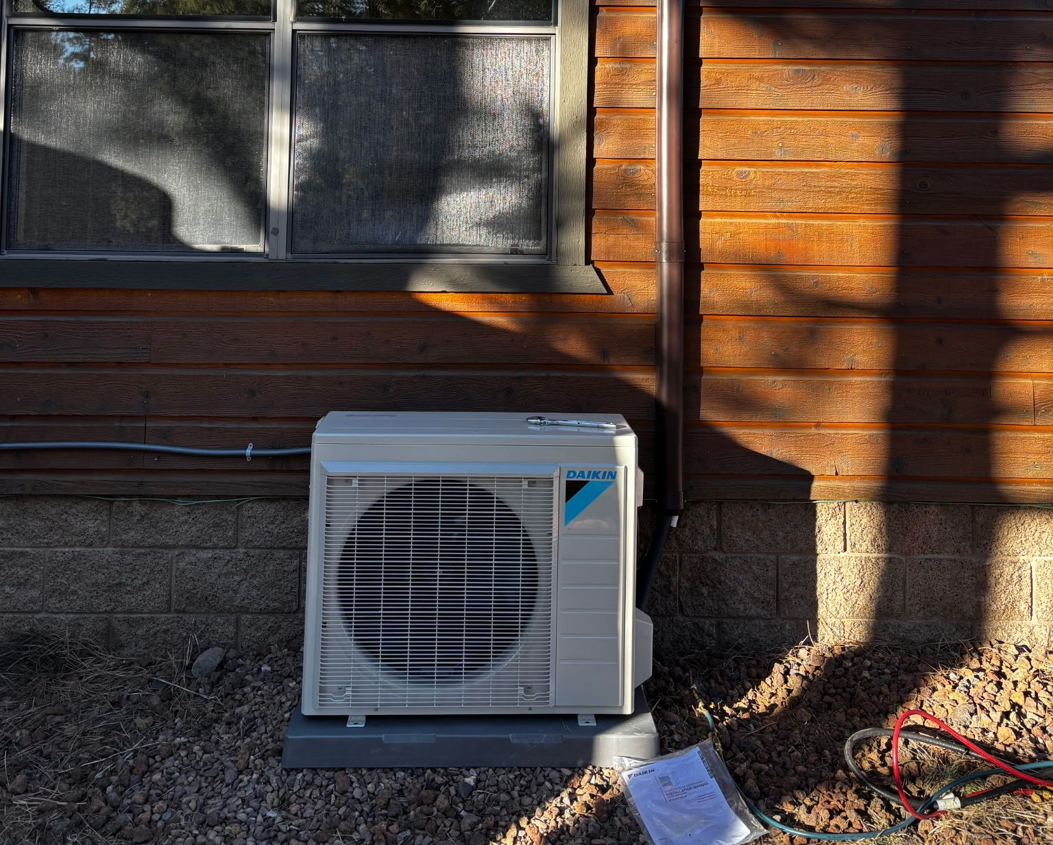 Outdoor wall-mounted AC unit beside a wooden house in sunlight.
