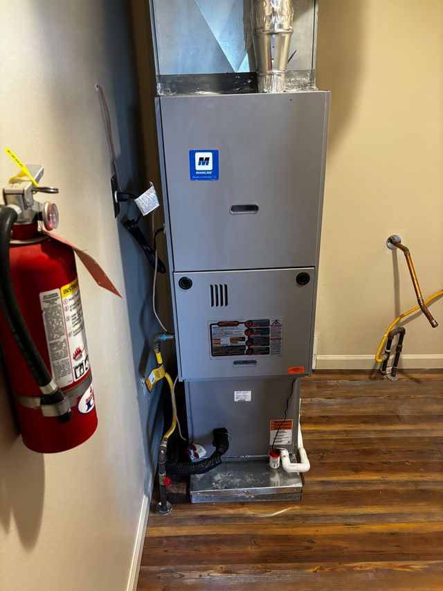 Gray HVAC furnace in a room with exposed ductwork, next to a red fire extinguisher on a wooden floor.