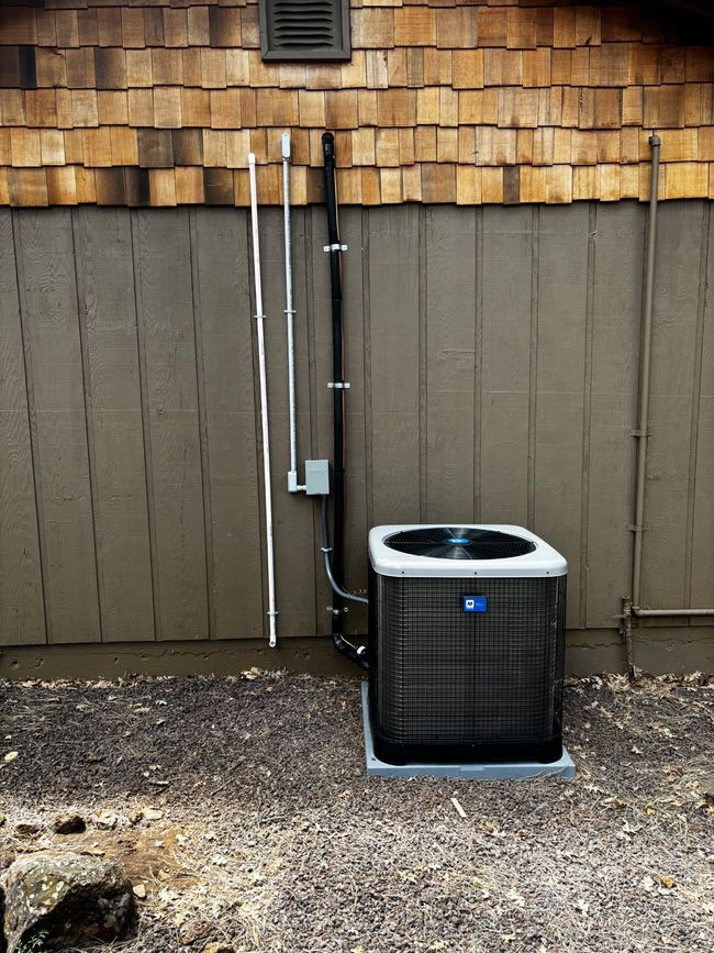 Outdoor HVAC unit beside a tan house wall with exposed white and black utility pipes above gravel.