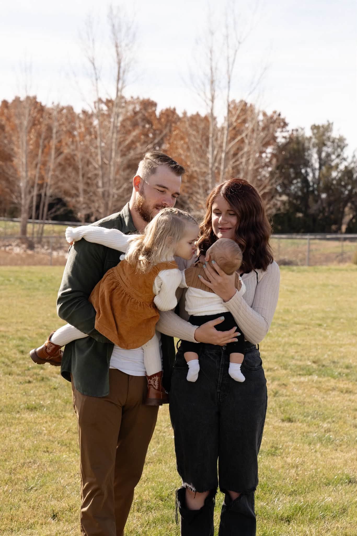 Two adults outdoors in a field holding two babies, with autumn trees in the background.