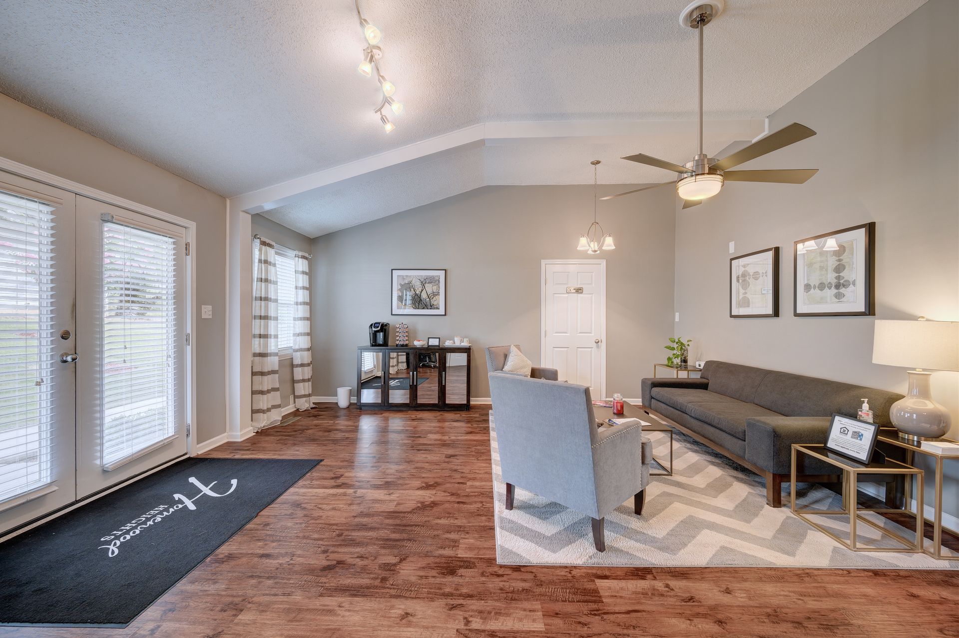 A modern living room with a gray sofa, armchair, patterned rug, wood flooring, and a ceiling fan.