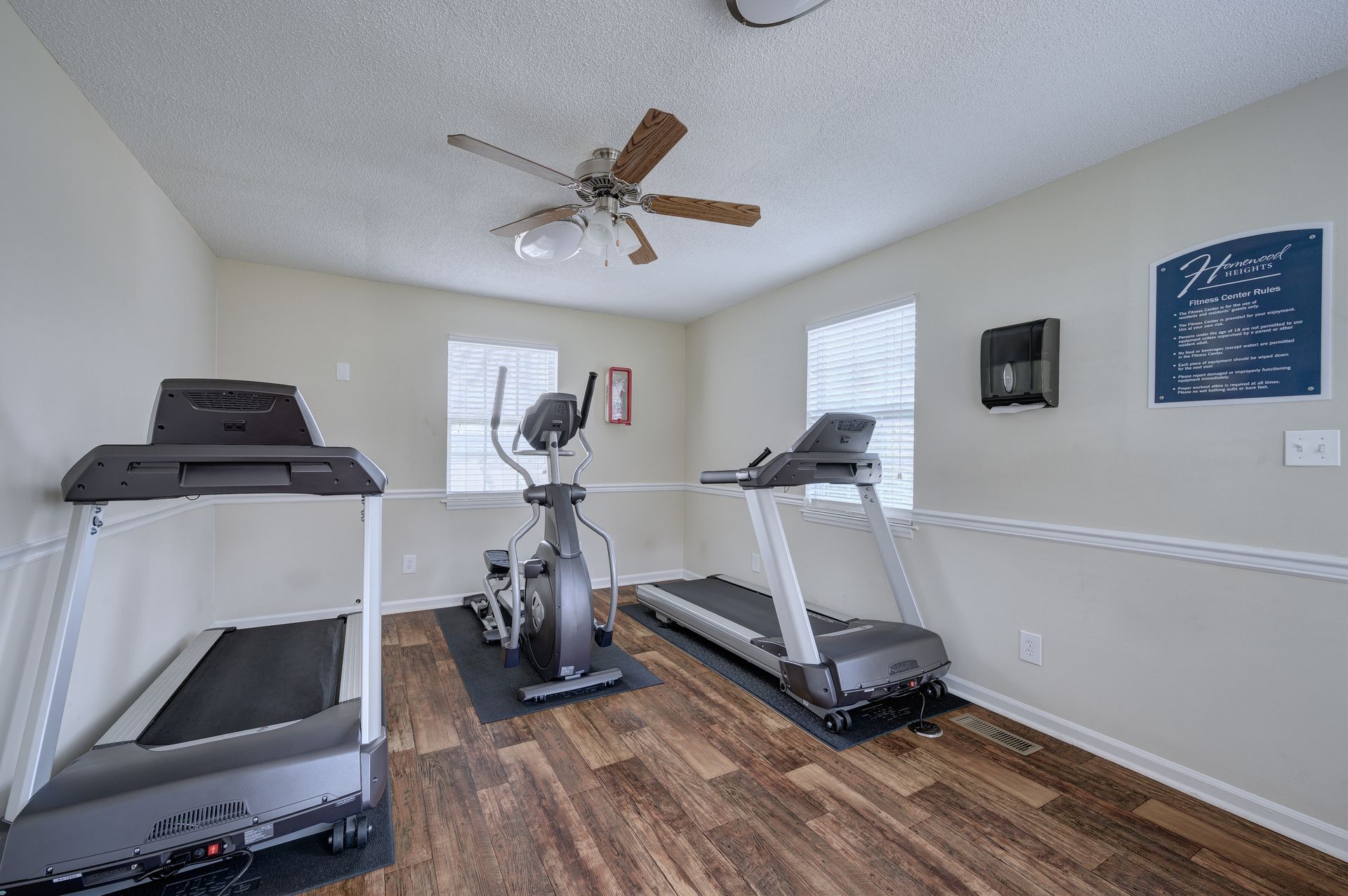 A gym with two treadmills on either side of an elliptical machine, set on wood-look flooring with light-colored walls.