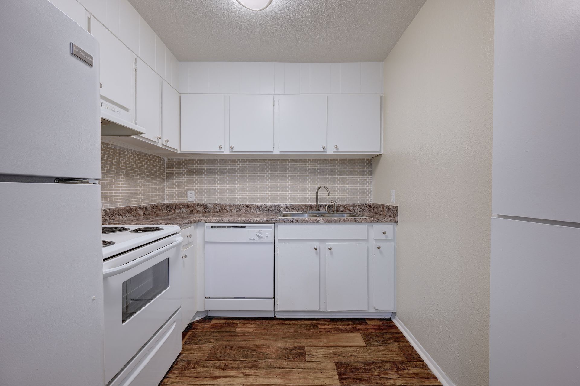 A small, galley-style kitchen with white cabinets, white appliances, speckled countertops, and wood-patterned flooring.