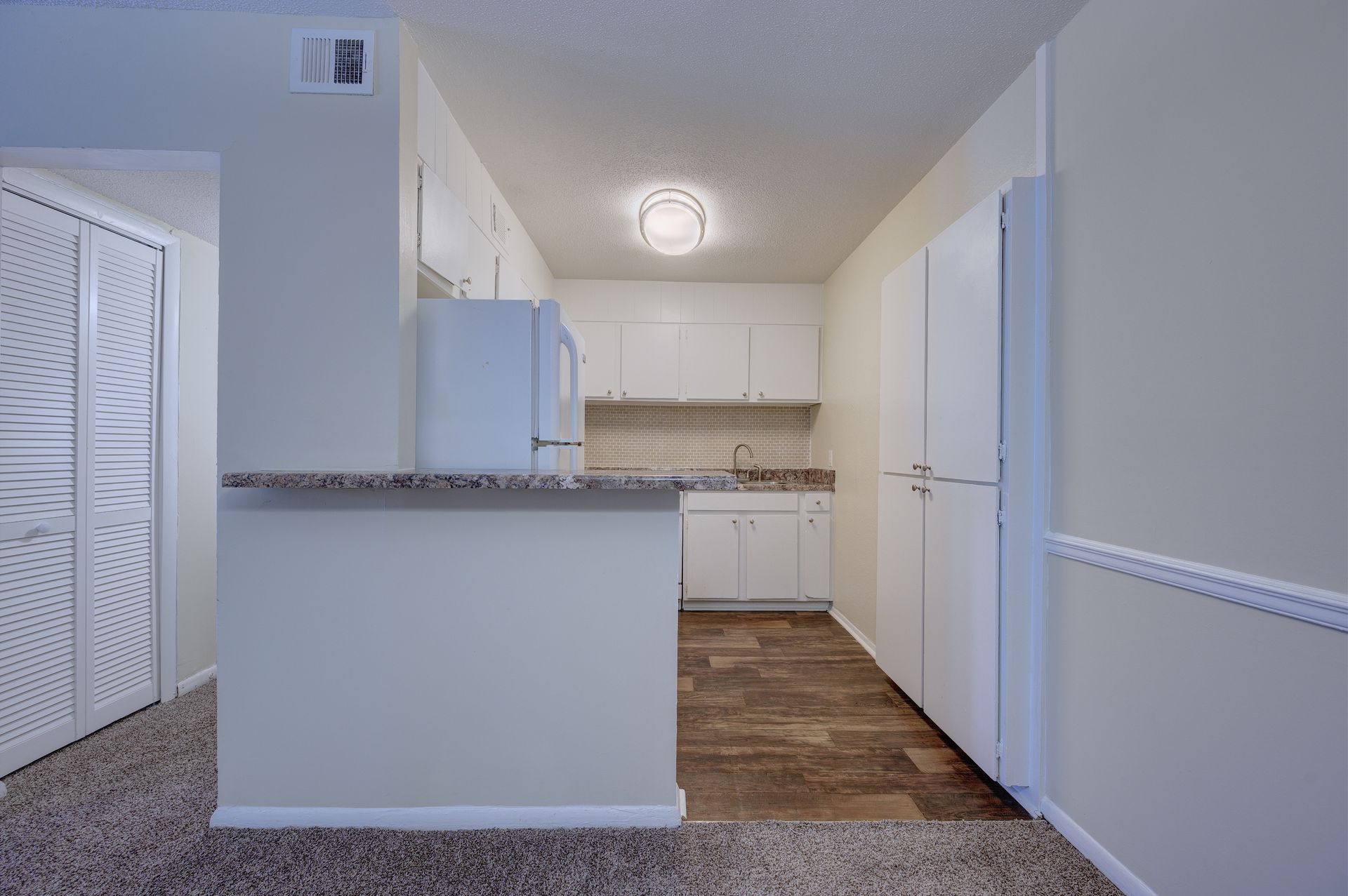 A kitchen with white cabinets, a refrigerator, and a small counter opening, viewed from a carpeted area.