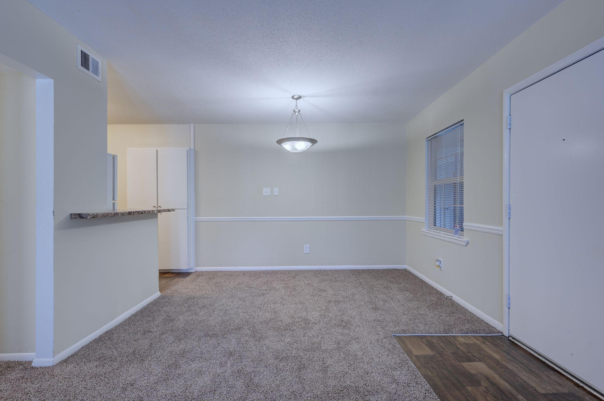 An empty dining area with beige walls, gray carpeting, a hanging light fixture, and a white door on the right.