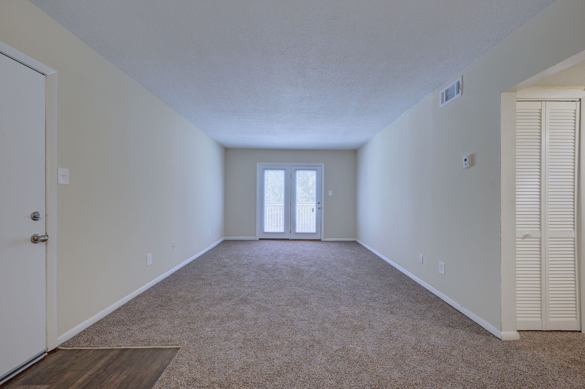 An empty room with light walls and beige carpet, featuring a white door, a closet with louvered doors, and glass patio doors.
