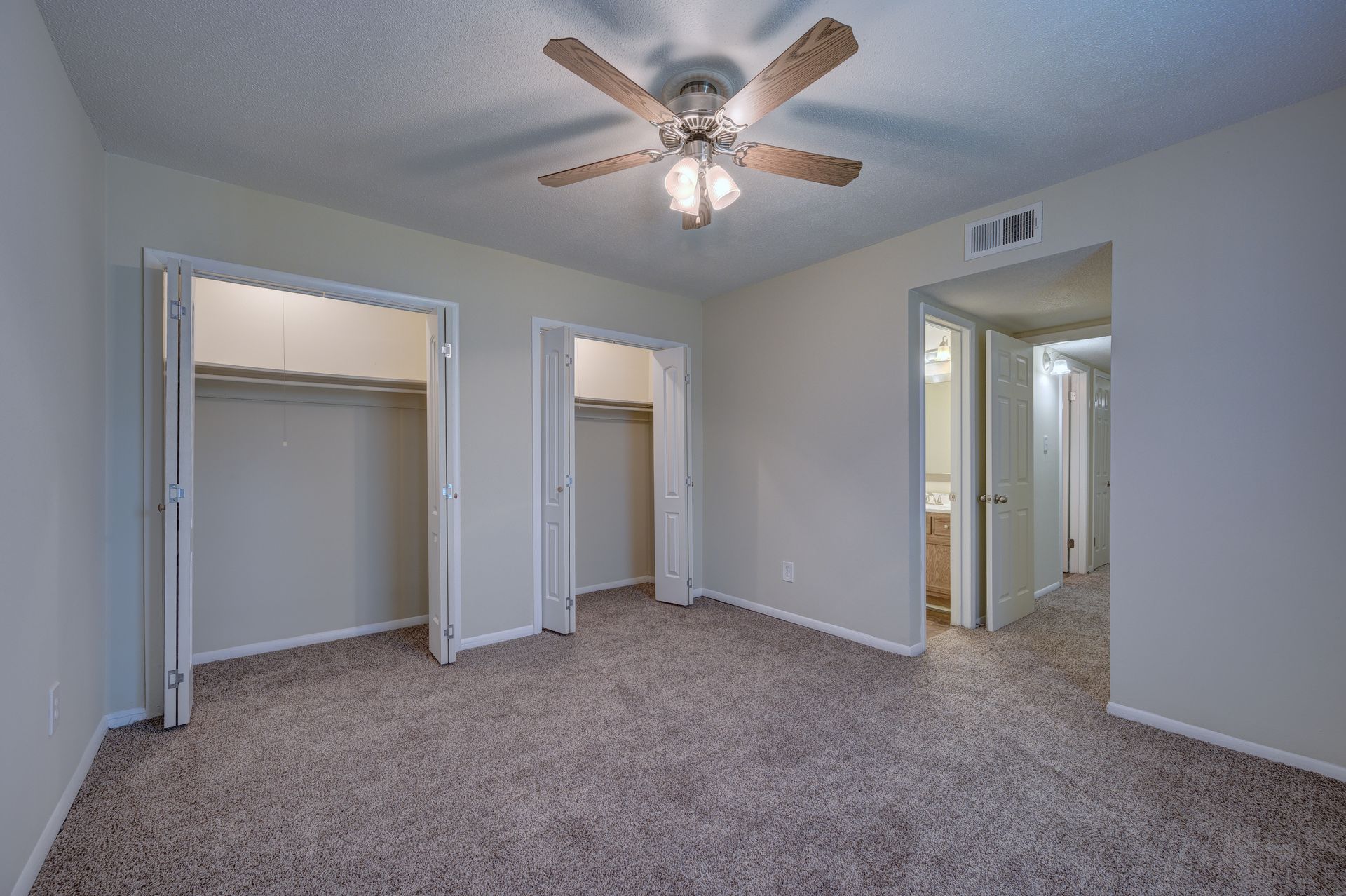 An empty bedroom with beige walls, patterned carpet, a ceiling fan, two closet doors, and an open doorway to a bathroom.