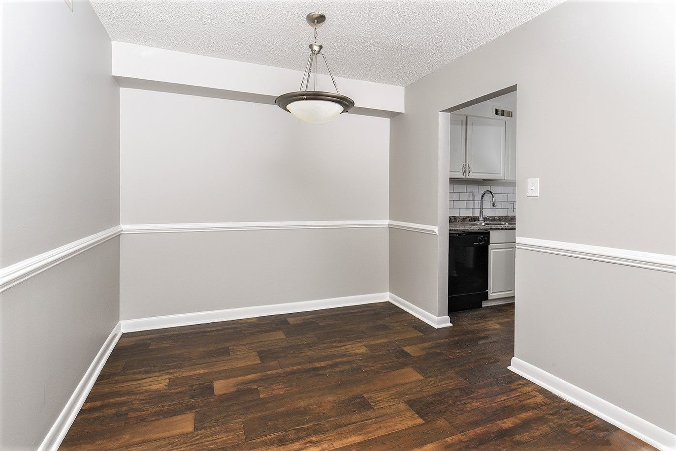 A dining area with gray walls, white wainscoting, a textured ceiling, a pendant light, and dark wood-look plank flooring.