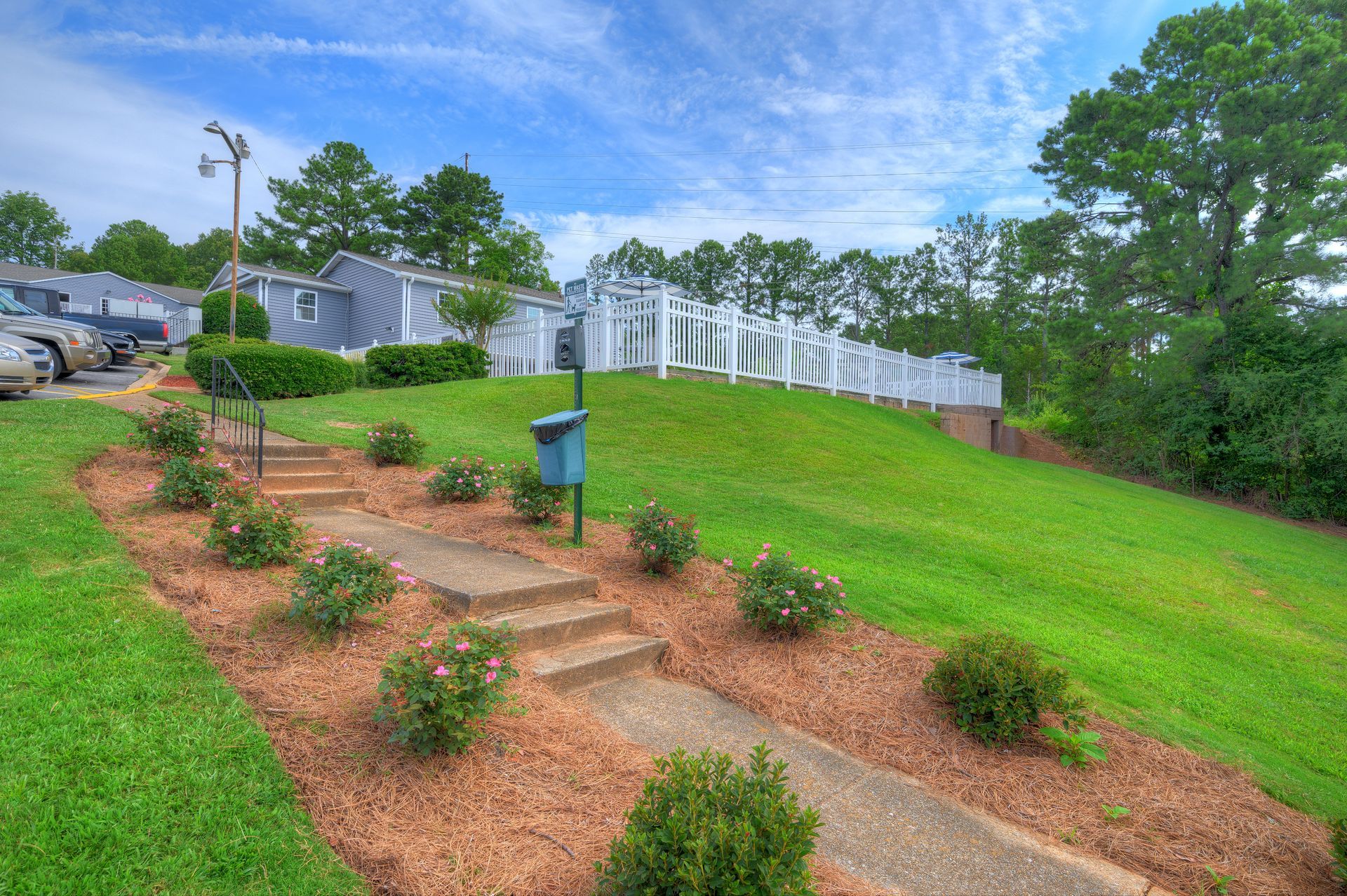 A stone pathway winds up a grassy hill toward a white fence, framed by mulched beds with small shrubs under a blue sky.