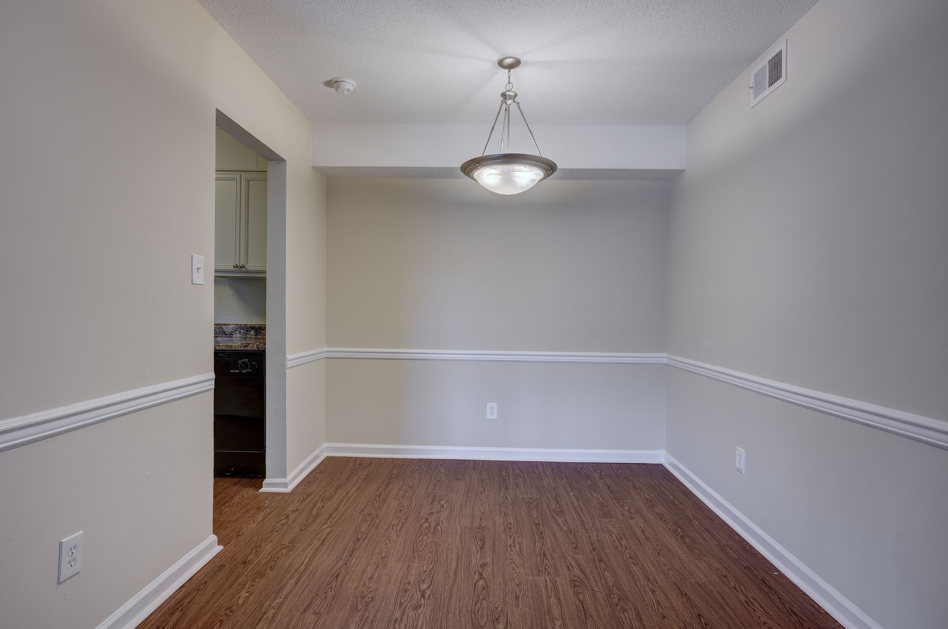 Empty dining area with wood-look flooring, light gray walls with chair rail molding, and a central pendant light fixture.
