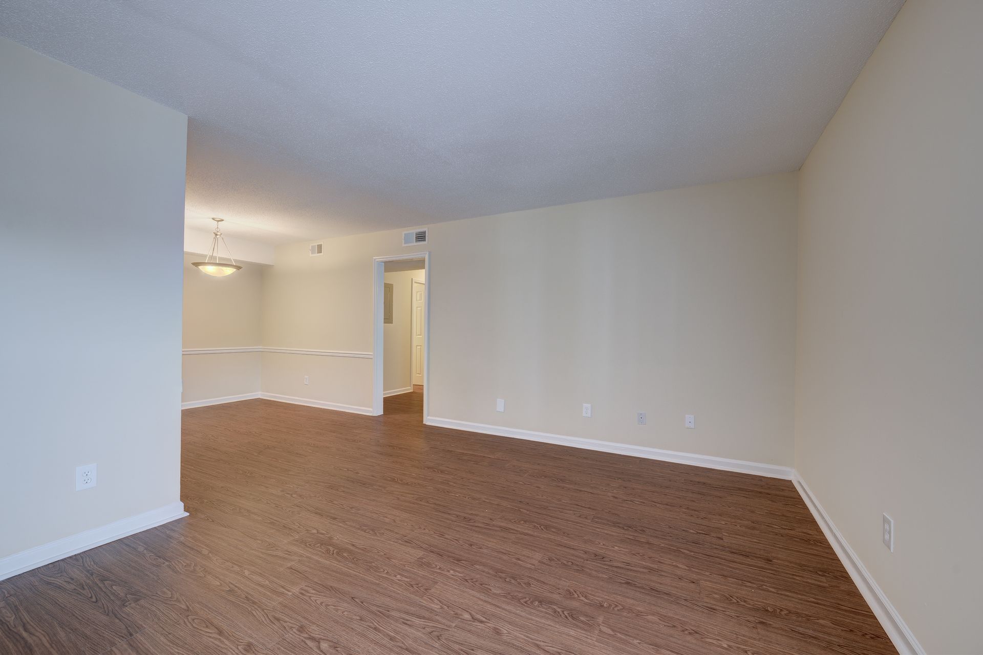 An unfurnished room with light beige walls and brown wood-look flooring leading to a dining area with a hanging light.