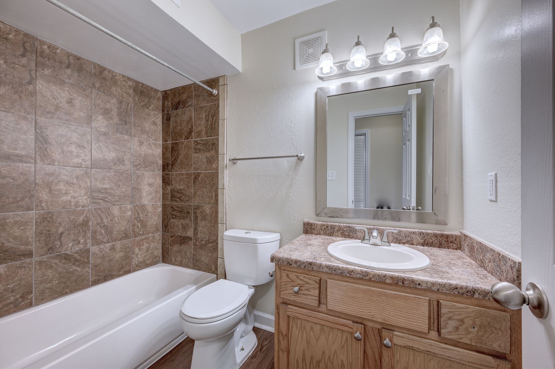 A bathroom with a white toilet, a wooden vanity with a stone countertop, a sink, a mirror, and a tiled shower-tub combo.