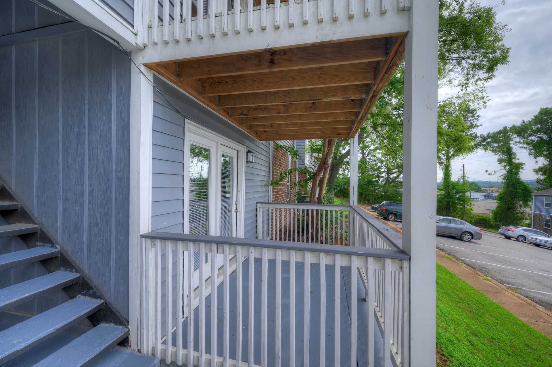 A blue porch with white railings and an open wooden ceiling under a second-story balcony next to outdoor stairs.