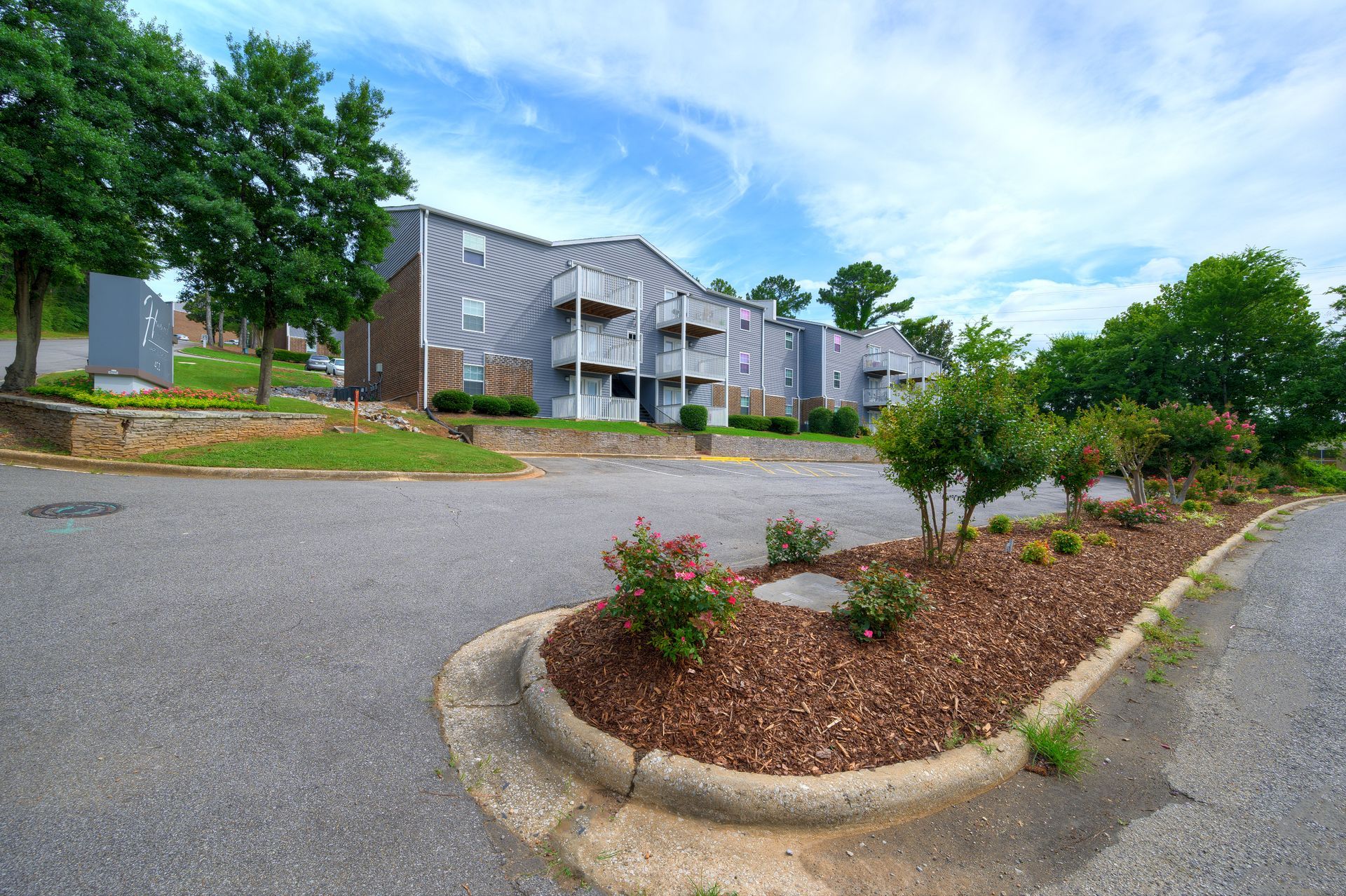 A multi-story apartment building with a gray facade, surrounded by trees, an asphalt parking lot, and a mulched garden.