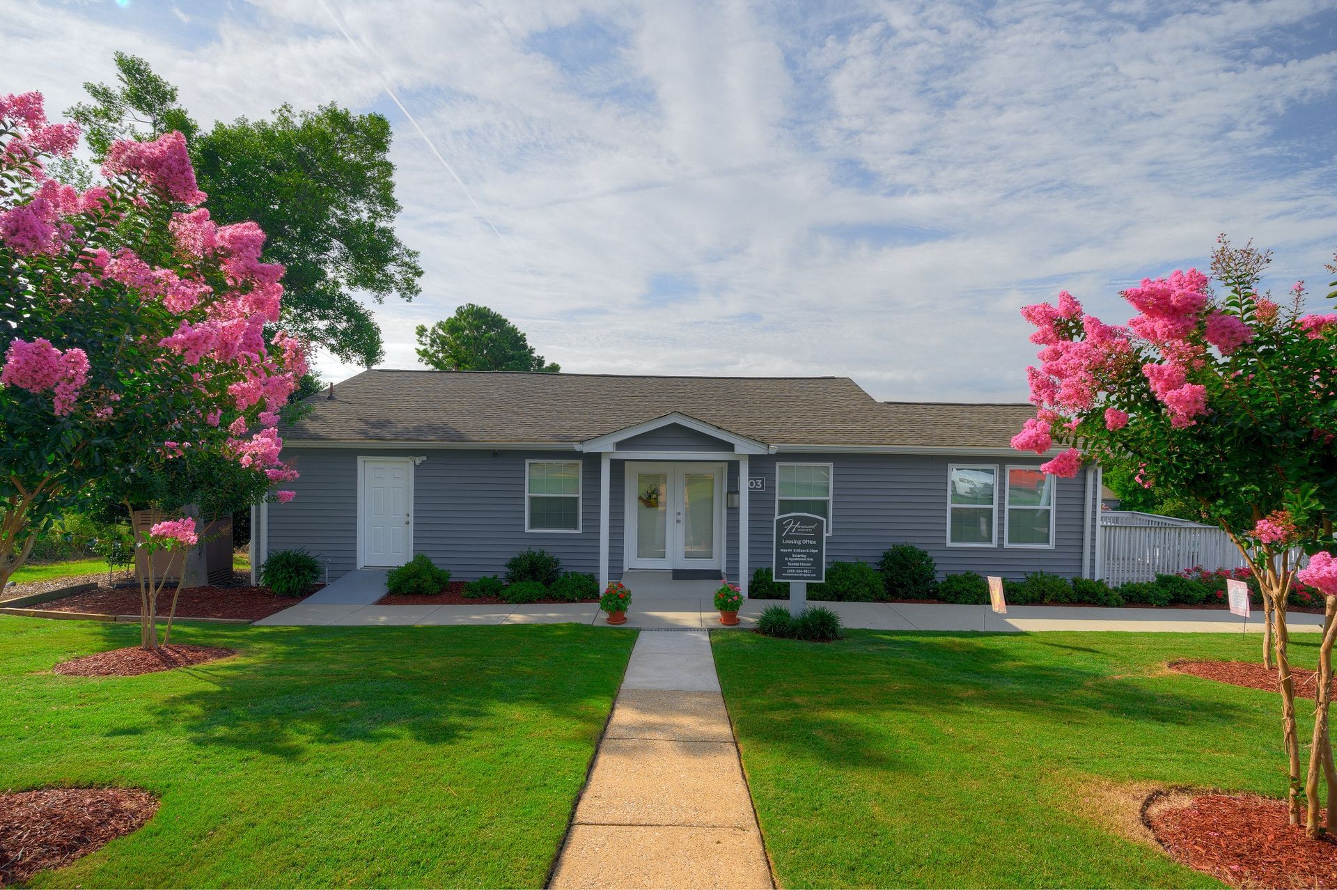 A single-story grey building with a white front door and walkway, framed by two large pink crape myrtle trees on a lawn.