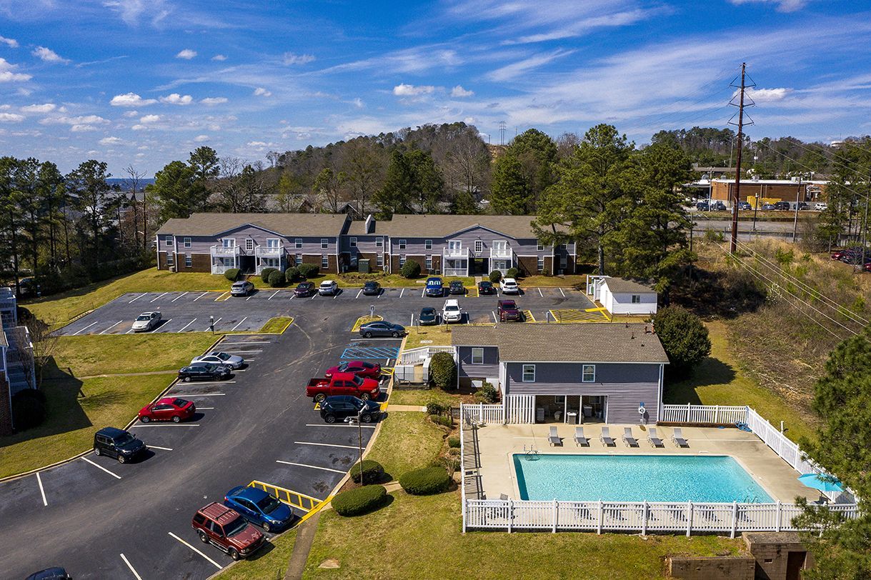 An aerial view of an apartment complex with a parking lot and a fenced-in swimming pool on a sunny day.
