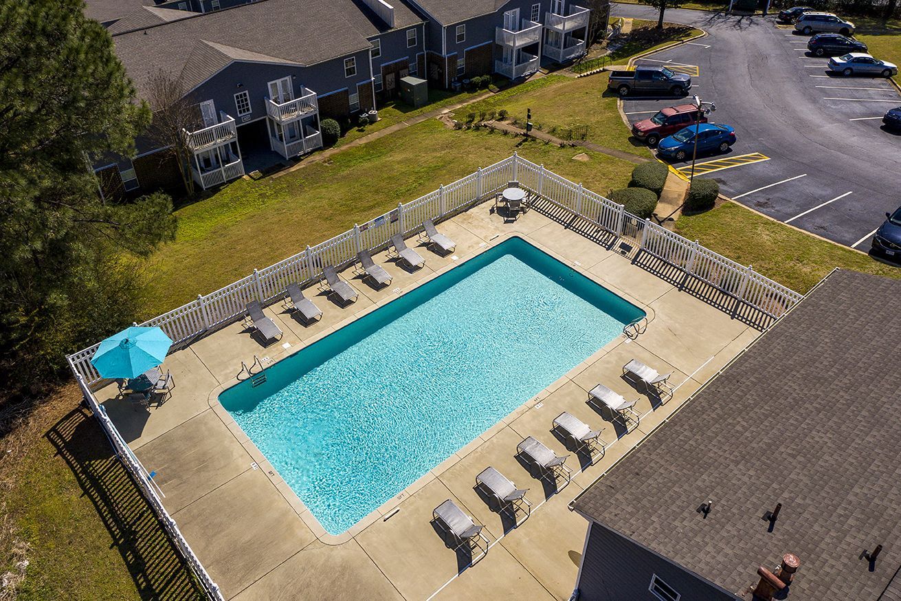 Aerial view of a rectangular apartment complex swimming pool surrounded by a fence, lounge chairs, and a parking lot.