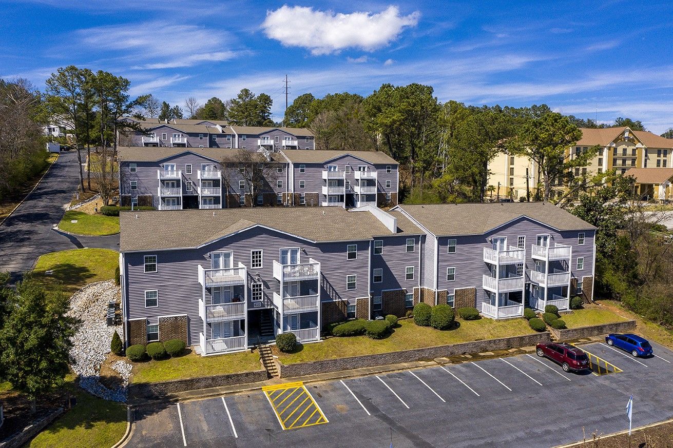 Aerial view of a gray apartment complex with white balconies, surrounding trees, and an asphalt parking lot.