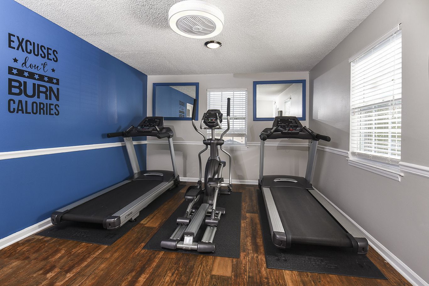 A small gym with a blue wall, two treadmills, one elliptical, hardwood floors, and windows with blinds.