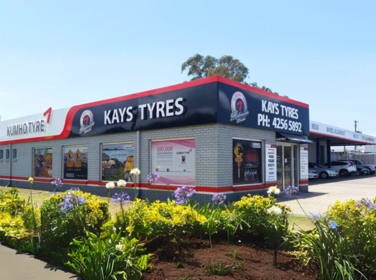 Kays Tyres storefront with black-red signage and flower beds in front under a bright blue sky