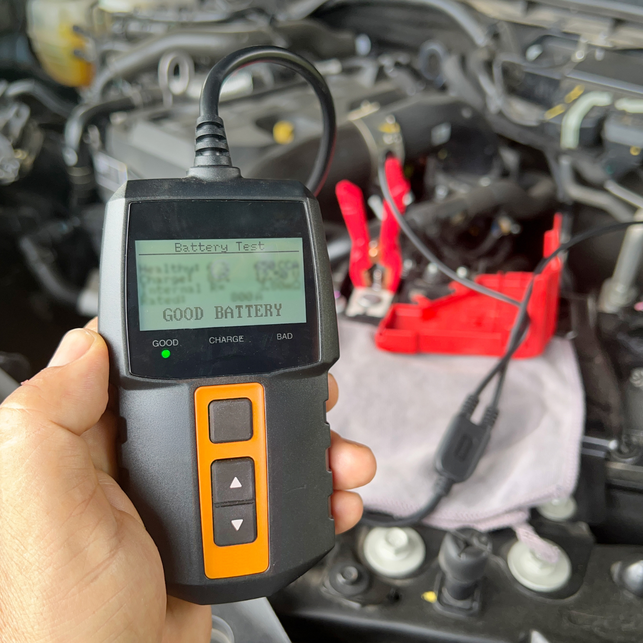 Handheld battery tester showing “GOOD BATTERY” in front of an open car engine, with red jumper cables nearby