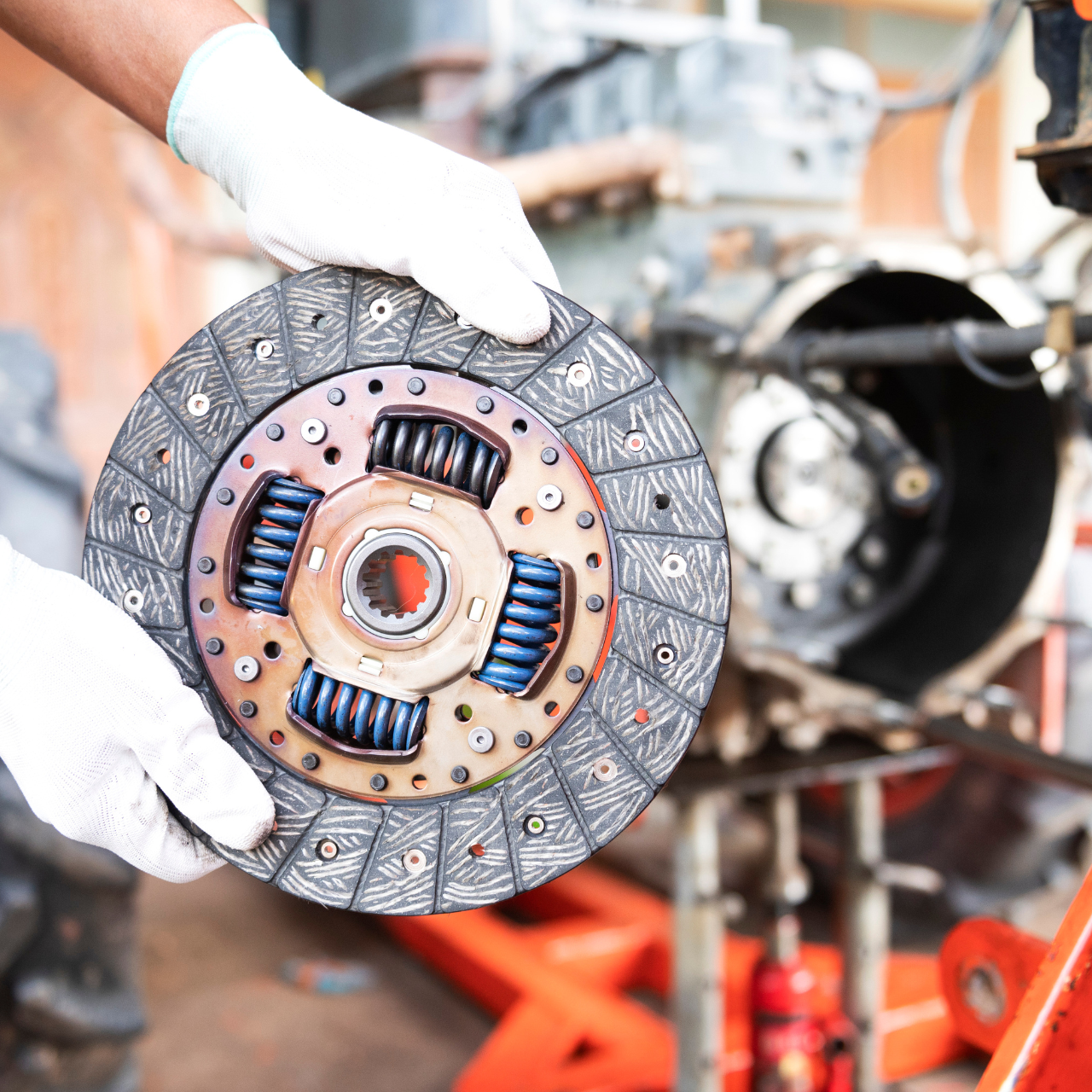Gloved hands holding a car clutch disc in an auto repair shop with a vehicle frame in the background