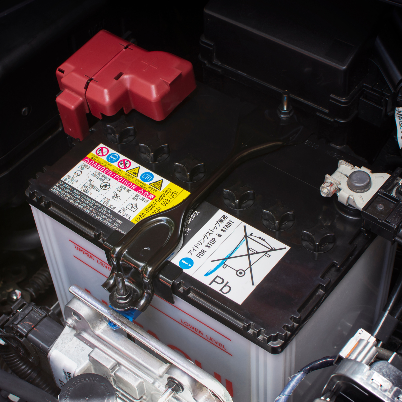 Car battery in a vehicle, with red positive terminal cover and warning labels visible.