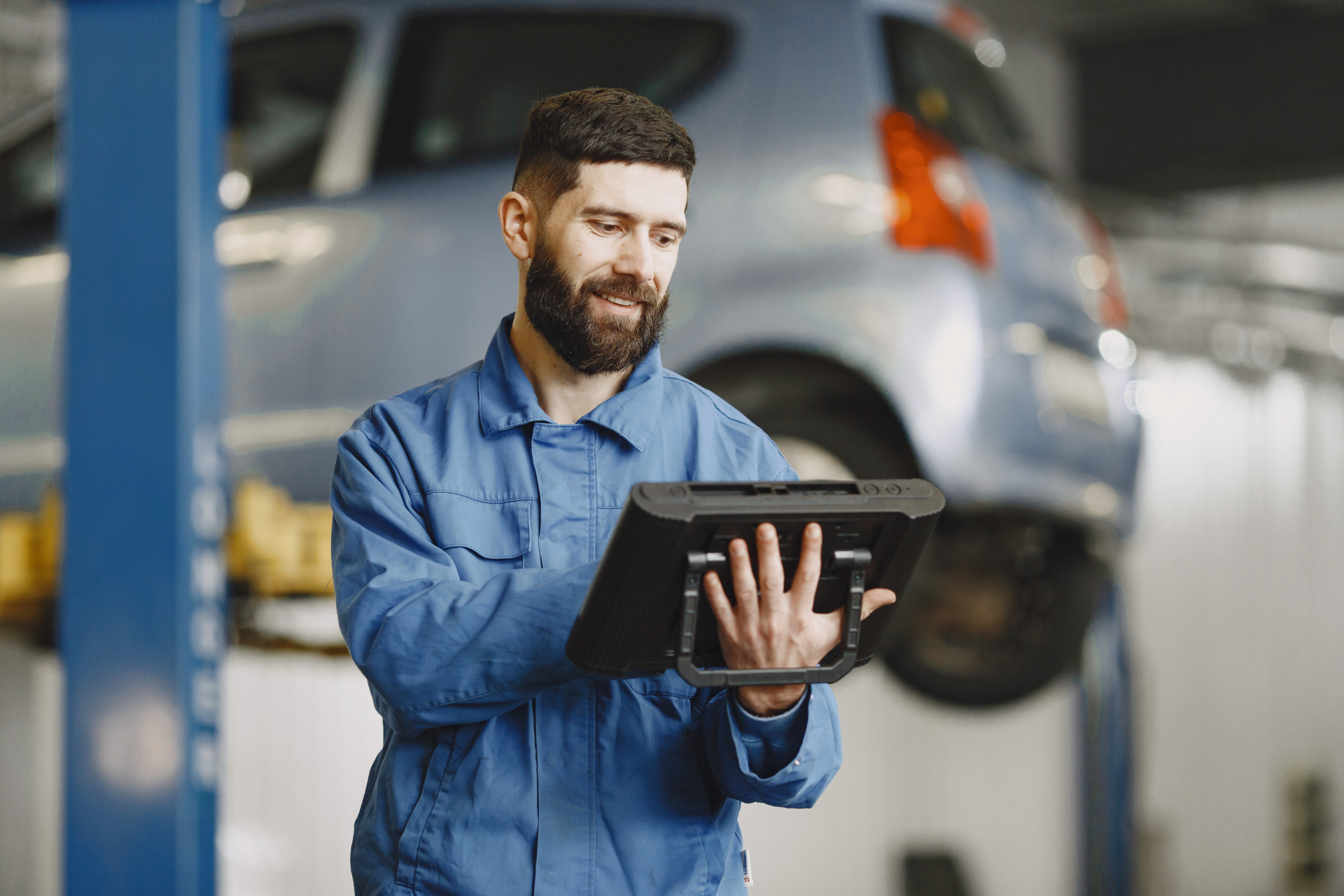 Mechanic in blue coveralls using a tablet in an auto repair garage with a raised car in the background