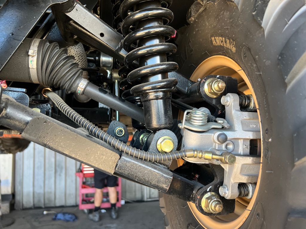 Close-up of a vehicle's front suspension system featuring a coil-over shock, control arm, brake caliper, and axle.