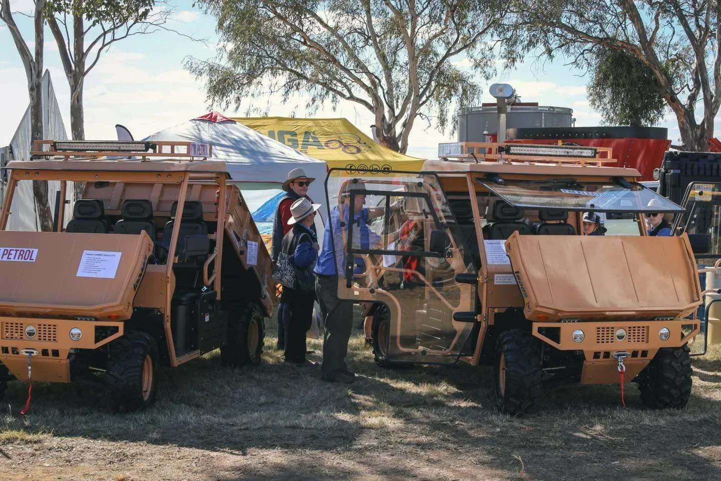 Two tan off-road utility vehicles parked outdoors under trees, with people standing near them.