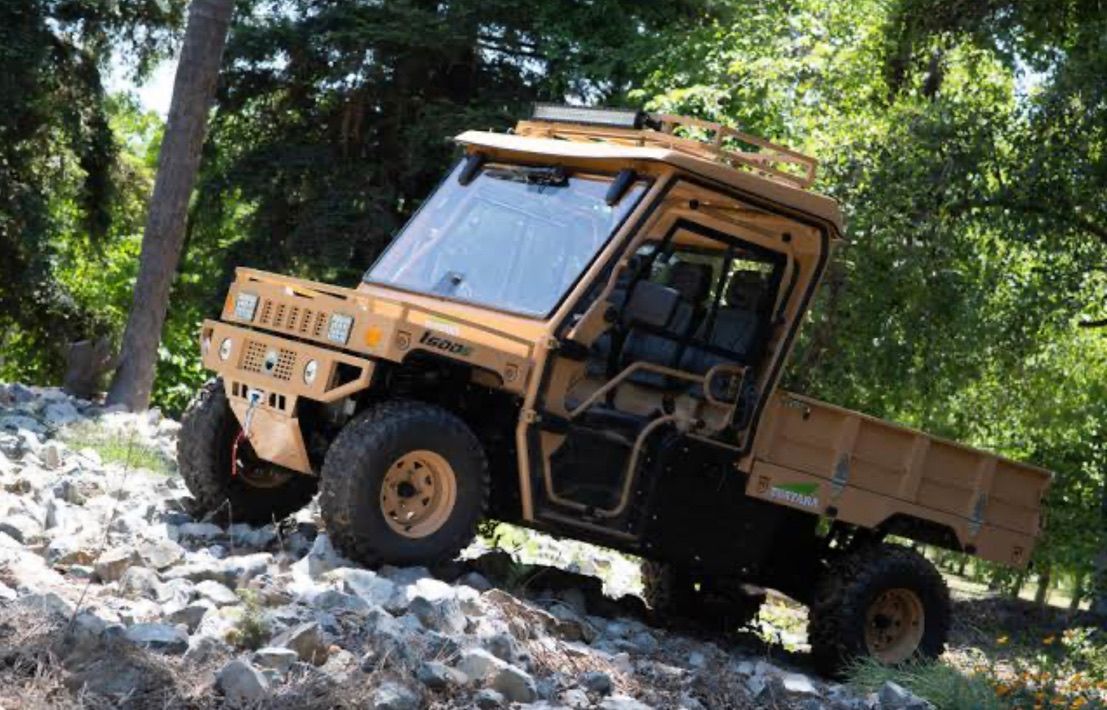 Tan utility vehicle traversing rocky terrain among trees.
