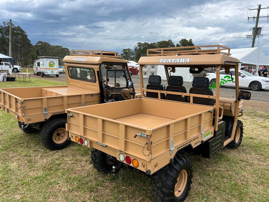 Two tan off-road utility vehicles with cargo beds parked on a grassy field under a cloudy sky.