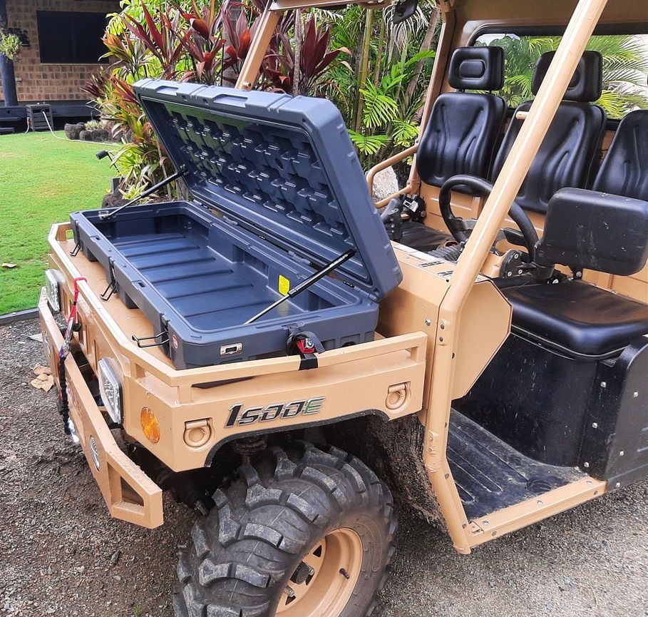 Tan off-road utility vehicle with an open front storage box, parked on a gravel driveway near greenery.