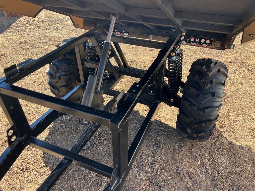 The undercarriage of a black, industrial dump trailer with two large, treaded tires and a central hydraulic lift cylinder.