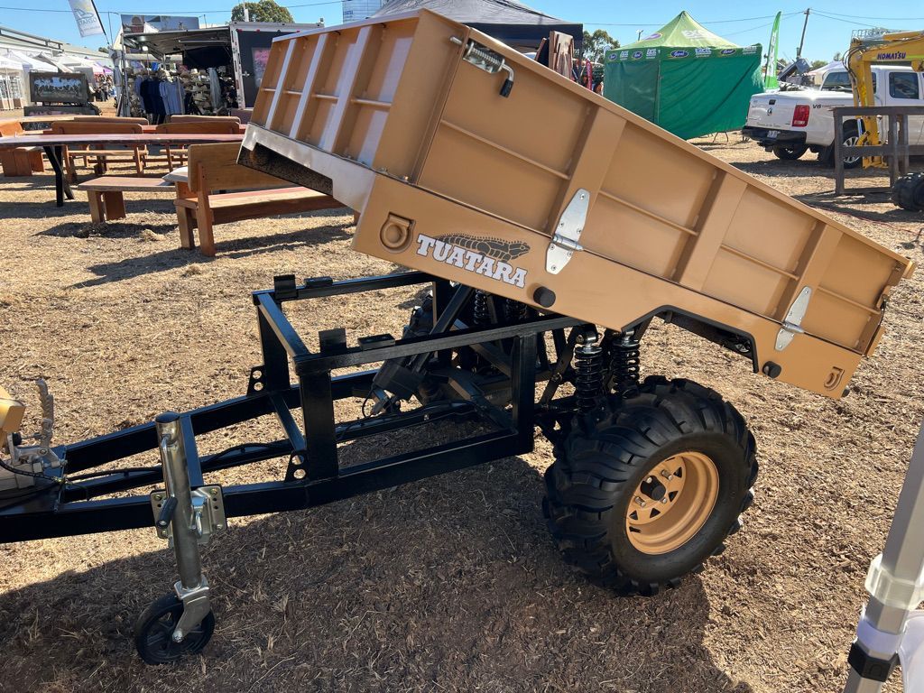 A tan, tilting utility trailer for ATVs sits on a wood-chipped outdoor display area under a bright, sunny sky.