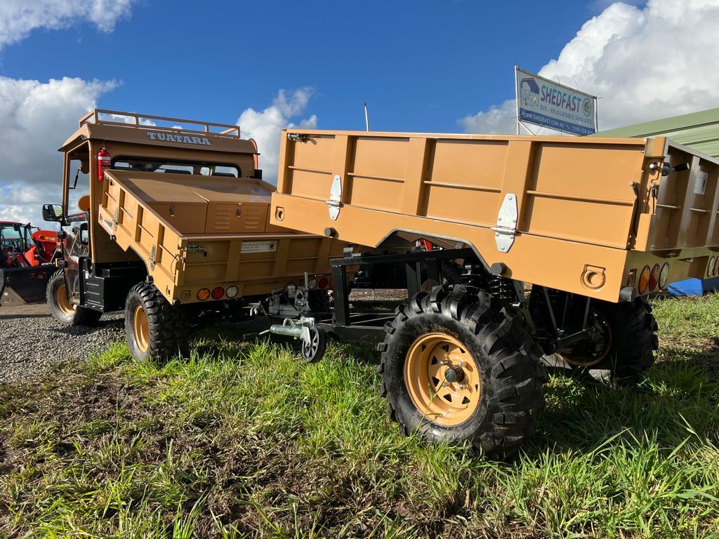 A tan-colored utility vehicle with a dump bed attached to a matching trailer, parked on a grassy field.