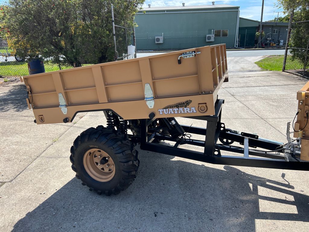 A tan-colored dump trailer with off-road tires, parked on a concrete surface outside a building.