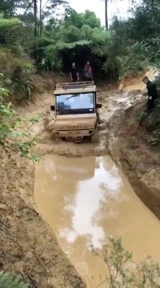 A tan off-road vehicle driving through a muddy forest trail, with two people standing nearby.