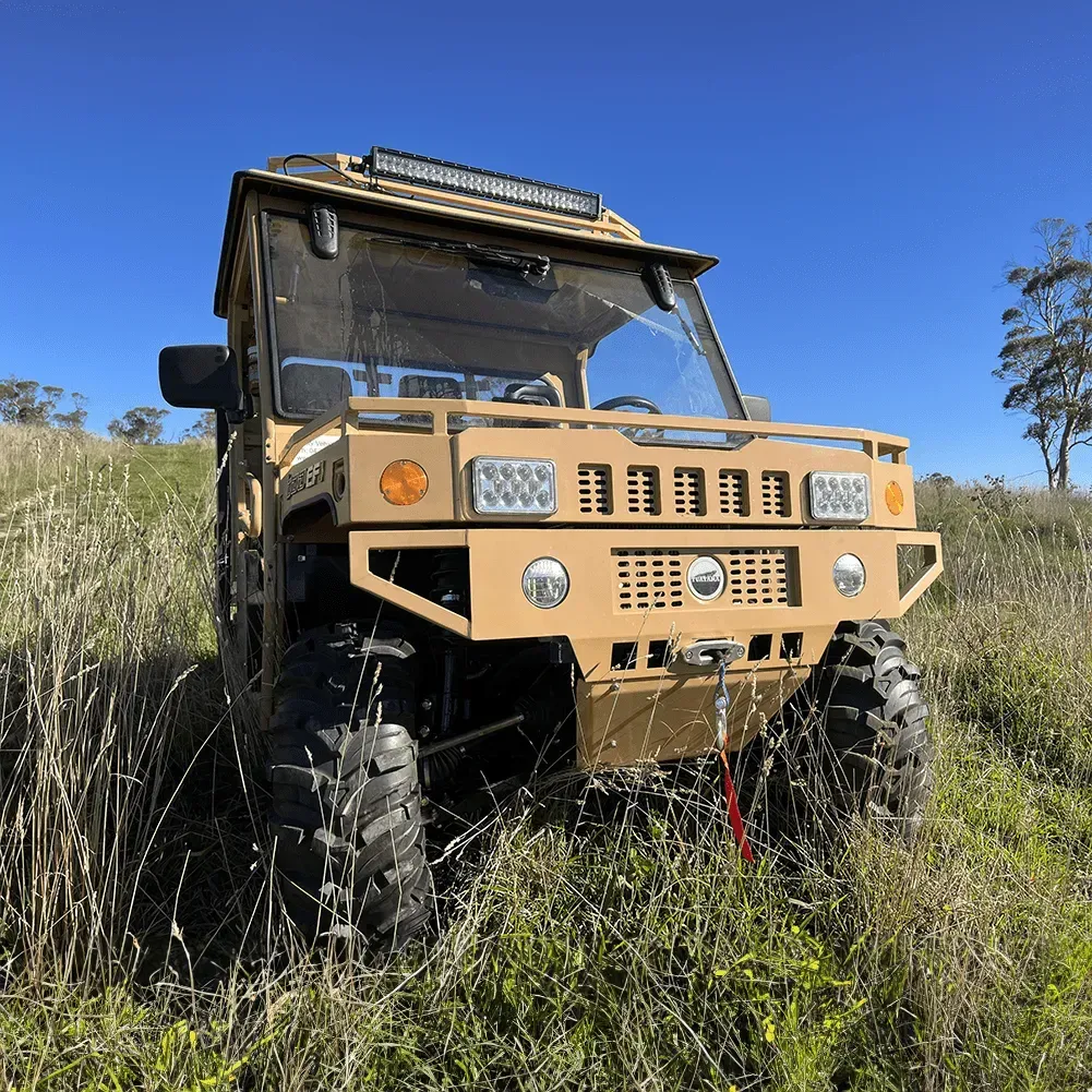 A tan, Humvee-style off-road vehicle sits parked in tall grass against a bright blue sky.