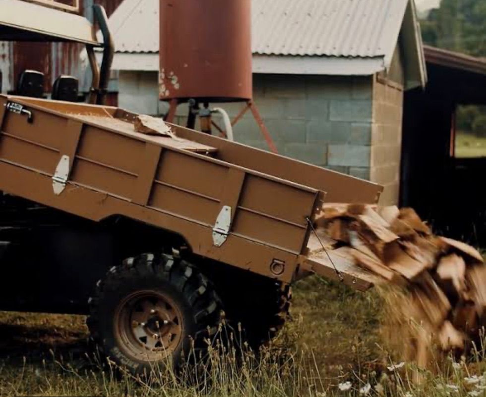 A tan utility vehicle dumping a load of firewood on grassy ground near a building.