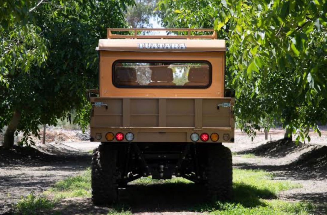 A tan, vintage-style utility vehicle with a covered bed sits centered on a dirt path between rows of green trees.