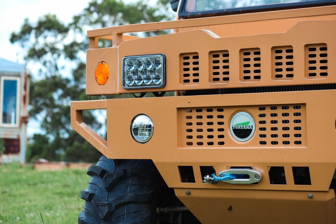 The front corner of a tan-colored off-road vehicle with a heavy-duty bumper, winch, and large, textured tire.