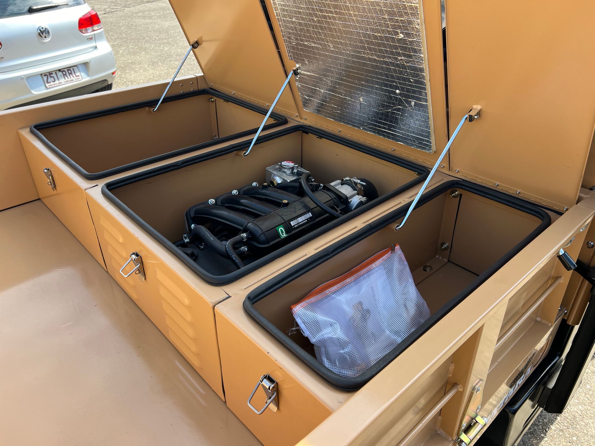 Three open tan storage compartments in a truck bed, one containing a car engine part and another a plastic bag.