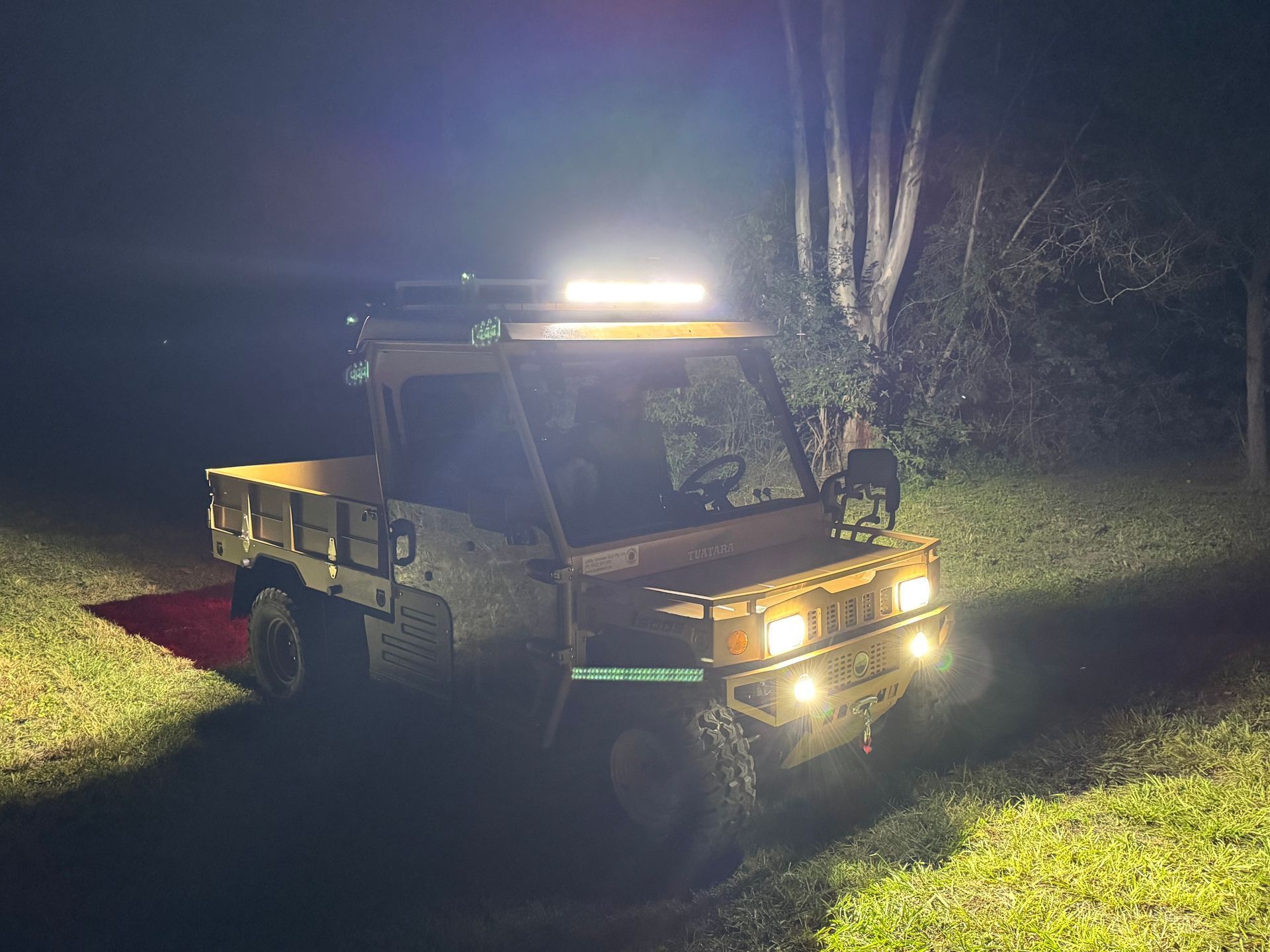 An off-road utility vehicle with illuminated headlights and a roof light bar sits on a grassy field at night.