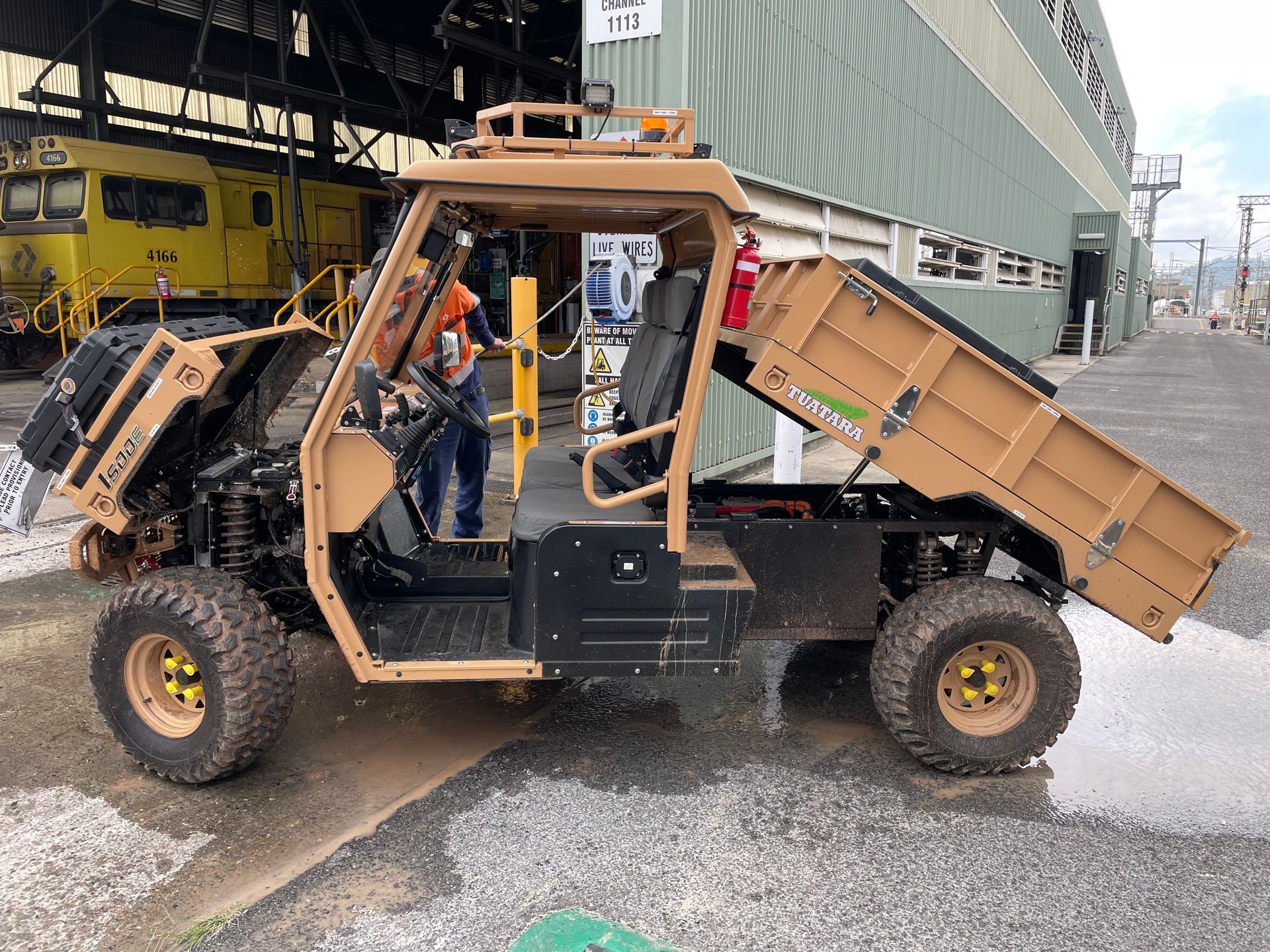 A tan utility task vehicle with a raised dump bed parked in an industrial yard next to a yellow locomotive.
