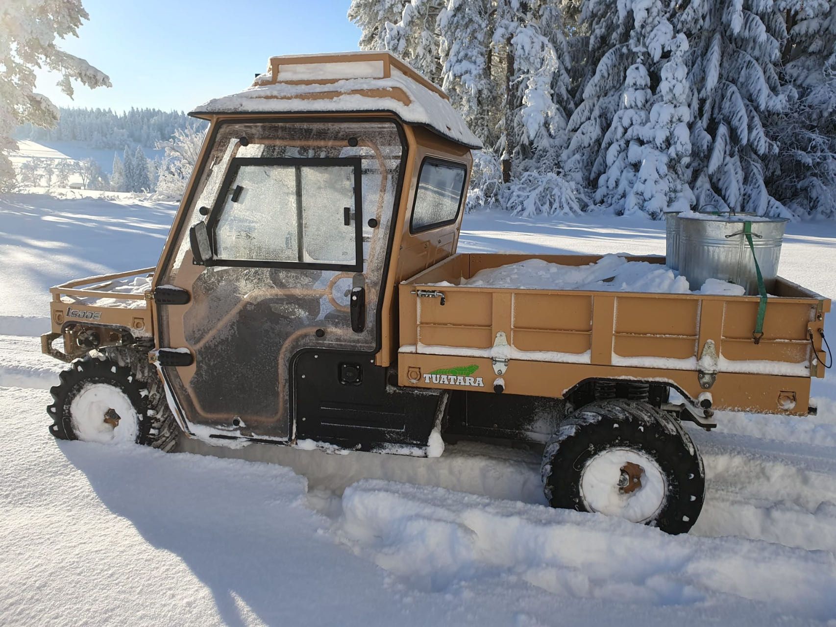 A tan utility vehicle with an enclosed cab and cargo bed parked in deep snow next to snow-covered pine trees.