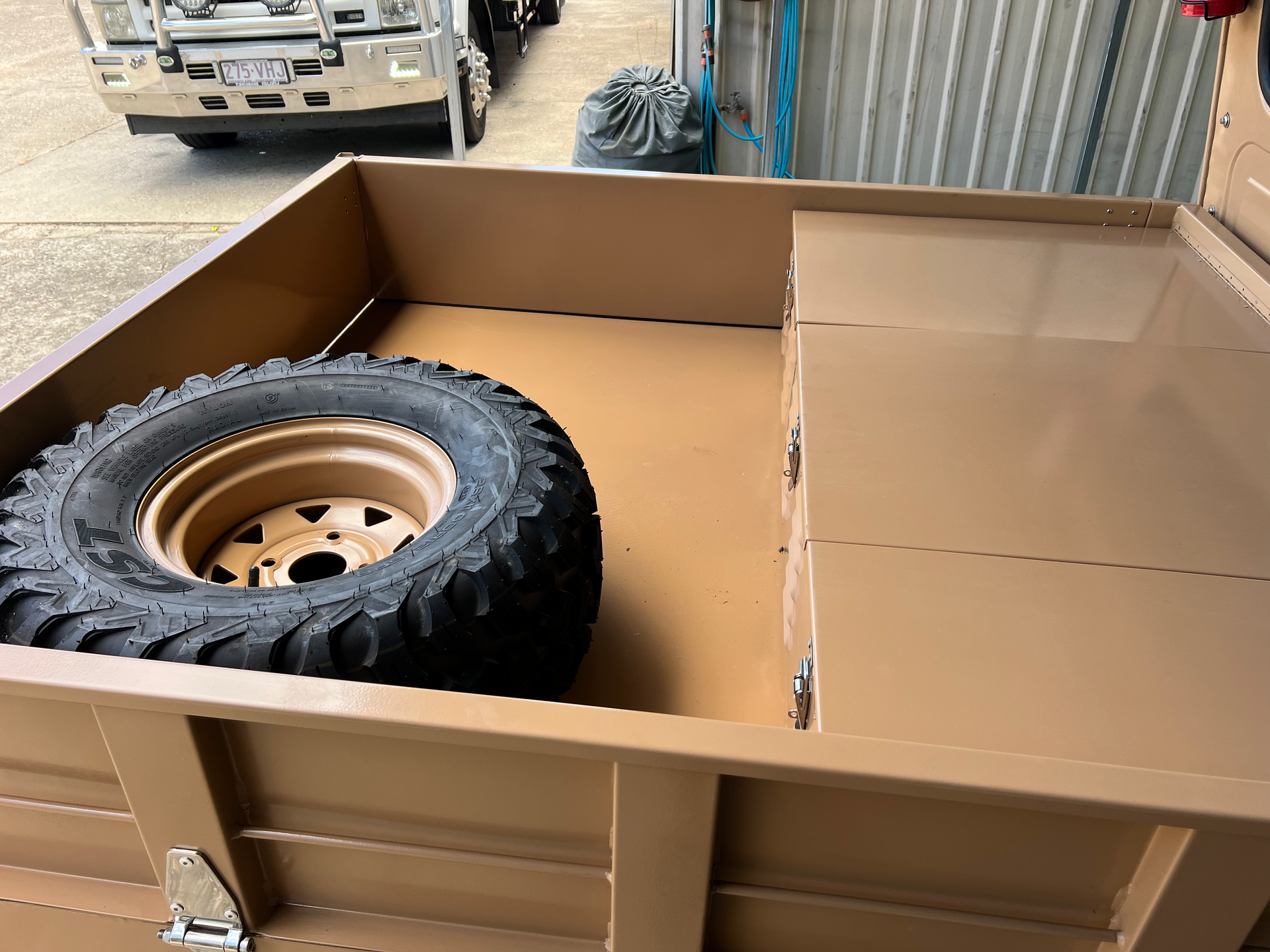 Tan metal trailer interior with a spare tire and a covered storage compartment, parked near a truck front.