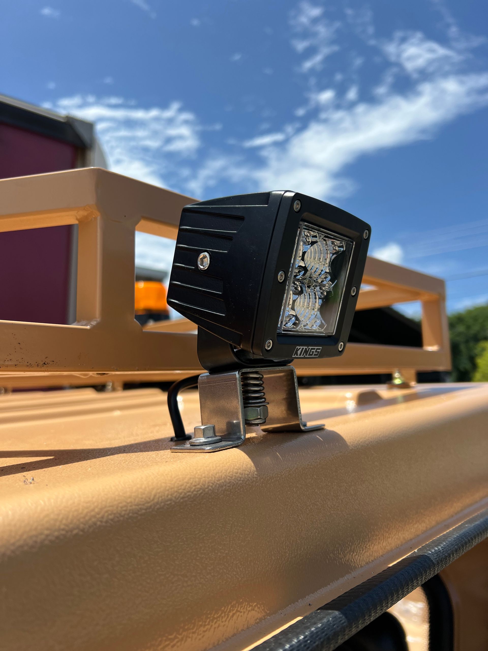 A black LED work light mounted to the beige roof of a vehicle with a metal storage rack against a bright blue sky.