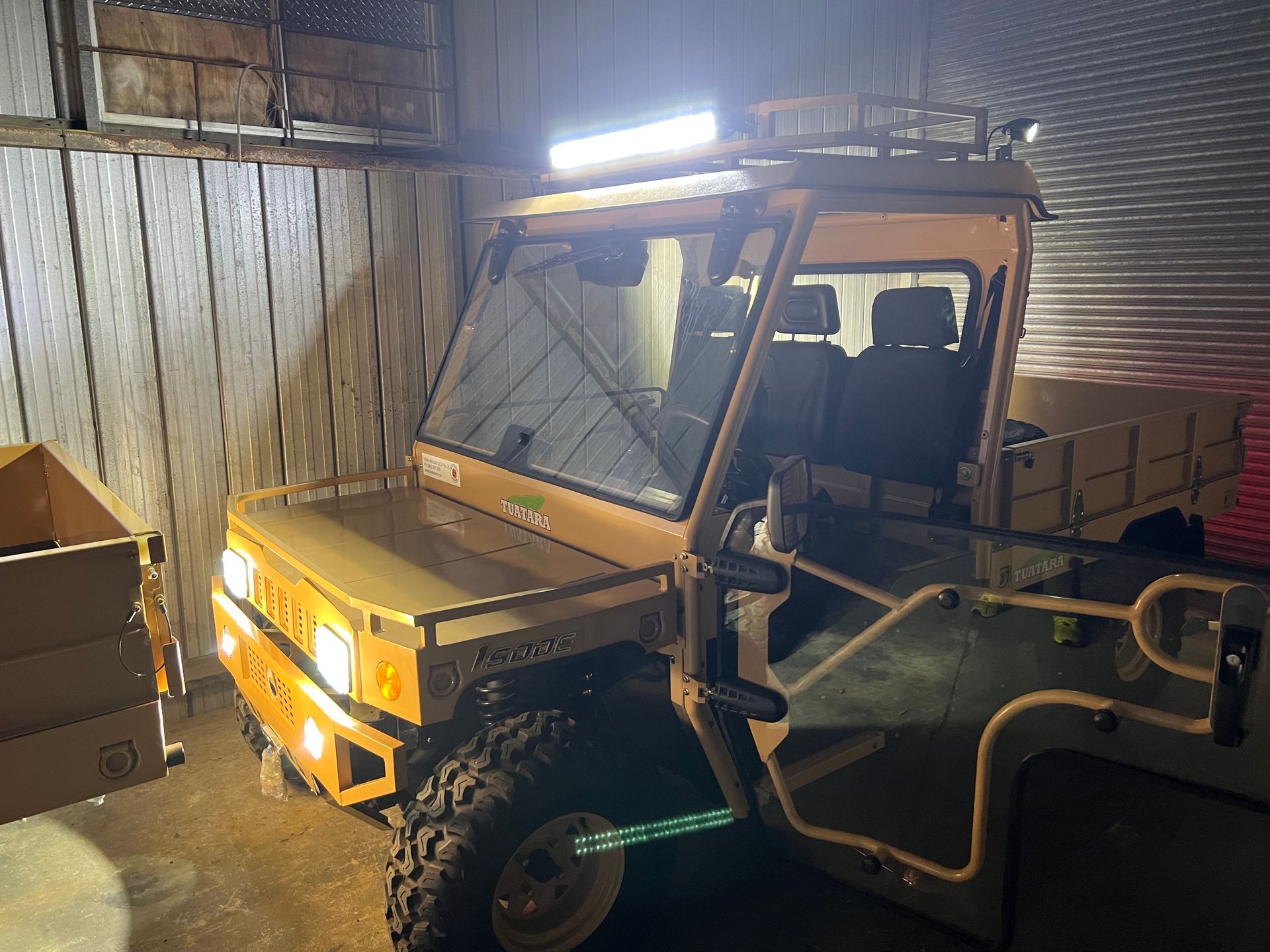 A tan, boxy utility vehicle with a roll cage and open doors parked inside a building with a metal wall.