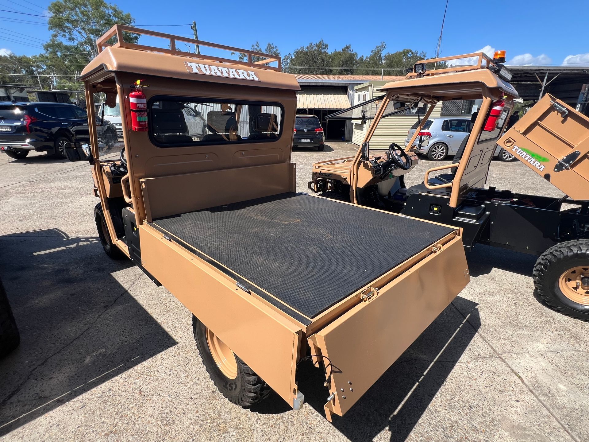 A tan-colored utility vehicle with a flatbed cargo area parked on a paved lot with other similar vehicles in the background.